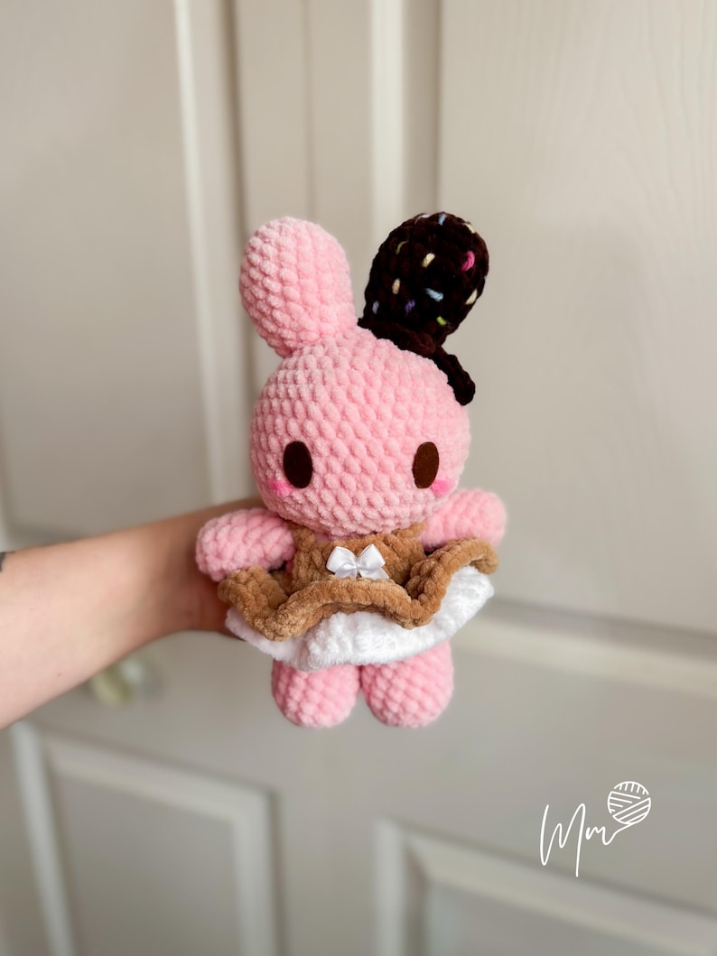 May include: A pink crocheted bunny plushie with brown eyes and a chocolate-covered ear. The bunny wears a brown and white dress with a small white bow. The plush toy is soft and cuddly, perfect for children.