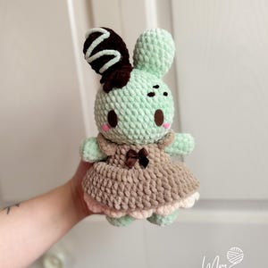 May include: A handmade crochet bunny doll in mint green, brown, and pink. The bunny has a brown and white swirl on its ear, a brown bow, and a dress with a pink trim. The doll is held in a person's hand.
