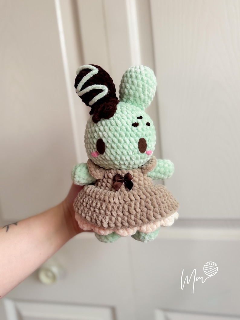 May include: A handmade crochet bunny doll in mint green, brown, and pink. The bunny has a brown and white swirl on its ear, a brown bow, and a dress with a pink trim. The doll is held in a person's hand.