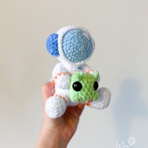 May include: A white crocheted astronaut with blue and orange details holding a green crocheted alien.