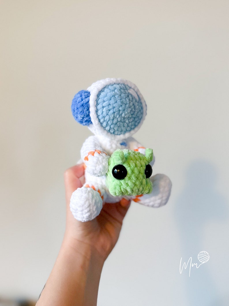 May include: A white crocheted astronaut with blue and orange details holding a green crocheted alien.