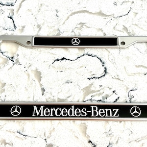 Puede incluir: Marco de matrícula plateado con detalles en negro. El marco presenta el logotipo de Mercedes-Benz y el nombre de la marca en letras blancas. El marco es rectangular y tiene orificios de montaje.