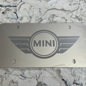 May include: A rectangular silver-toned metal license plate frame featuring the word MINI centered within a circular emblem with stylized wings on both sides. The plate has four mounting holes, one in each corner, and a smooth, reflective finish.