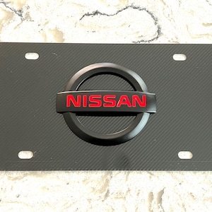 May include: Black license plate with a carbon fiber texture and the red "NISSAN" logo. The plate has four mounting holes and a black circular emblem. The plate is designed for a vehicle.