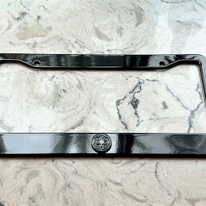Op de afbeelding: Een glanzend zwart kentekenplaatraam met een decoratief embleem in het midden. Het frame heeft een rechthoekige vorm met afgeronde hoeken en montagegaten. Het embleem is een cirkelvormig ontwerp met details.