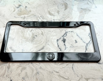 May include: A glossy black license plate frame with a decorative emblem at the bottom center. The frame has a rectangular shape with rounded corners and mounting holes. The emblem is a circular design with intricate details.