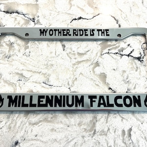 May include: A silver license plate frame with the text "MY OTHER RIDE IS THE" at the top and "MILLENNIUM FALCON" at the bottom in black. The frame has a shiny, metallic finish.
