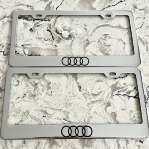 May include: Two chrome license plate frames with the Audi logo in black. The frames are rectangular with rounded corners and a polished finish. A small bag of hardware is visible between the frames. The background is a marble-like surface.