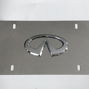 May include: A silver metal licence plate frame with a chrome Infiniti logo in the centre. The frame has four holes for mounting.