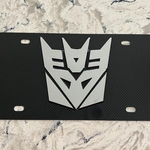 Peut inclure: Plaque d'immatriculation noire avec un emblème argenté des Decepticons de Transformers. La plaque présente une texture en fibre de carbone et quatre trous de montage.