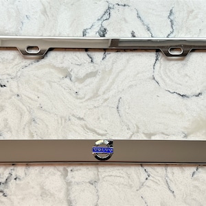 May include: Chrome license plate frame with a silver Volvo logo in the center. The logo has a blue background and a silver ring around it.