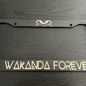 May include: Black metal licence plate frame with a silver Wakanda Forever inscription and a silver silhouette of a Black Panther mask. The frame also features a silver silhouette of two crossed arms.
