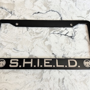 May include: Black license plate frame with the letters "S.H.I.E.L.D." in silver. The frame has the S.H.I.E.L.D. logo on each side. The frame is rectangular with rounded corners and mounting holes.