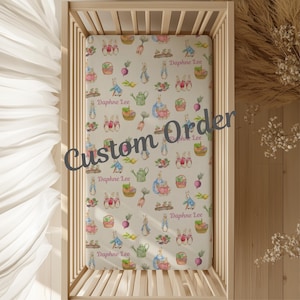 May include: A fitted crib sheet with a repeating pattern of bunnies, baskets, watering cans, and vegetables in pastel colors. The sheet is personalized with the name "Daphne Lee". The words "Custom Order" are printed across the sheet.