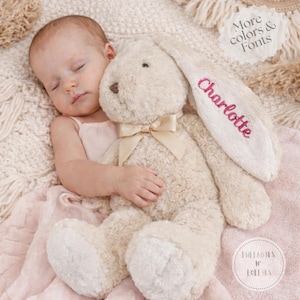 May include: A plush, cream-colored bunny rabbit with a bow tie and the name "Charlotte" embroidered in pink on one ear. The bunny is lying on a pink blanket. The image also includes the text "More colors & Fonts" and "Lullabies N' Lollies".