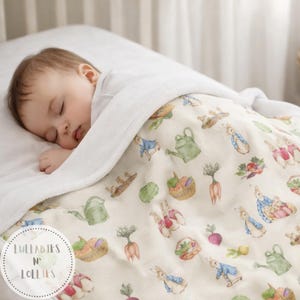 Puede incluir: Un bebé dormido bajo una manta blanca. La manta presenta un patrón caprichoso de conejos, regaderas y verduras en tonos verdes, azules y naranjas. La manta tiene el texto "Lullabies N' Lollies" en un círculo.