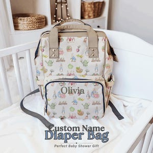 May include: A beige diaper bag featuring a pattern of rabbits, watering cans, and vegetables. It has a top handle, adjustable straps, and a front pocket with the name "Olivia". The text "Custom Name Diaper Bag" is printed at the bottom.