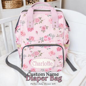 May include: Pink floral diaper bag with a rose pattern, featuring the name "Charlotte" in a white oval. The bag has a top handle, shoulder straps, and multiple pockets. Text on the bag reads "Custom Name Diaper Bag" and "Perfect Baby Shower Gift."