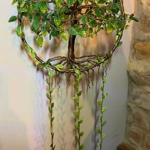 Op de afbeelding: Een handgemaakte wanddecoratie met een draadboomontwerp, groene bladeren en hangende ranken. De boom is geplaatst in een cirkelvormig frame. De bladeren hebben gele accenten. Het stuk is op een neutrale muur gemonteerd.