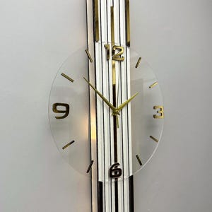 May include: A wall clock featuring a vertical rectangular base with white and black stripes and gold trim. A circular transparent face is mounted on the front, displaying gold numerals at 12, 3, 6, and 9, along with gold clock hands.