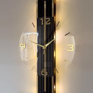 Puede incluir: Un reloj de pared con un panel rectangular vertical negro, borde dorado e iluminación trasera integrada. Una esfera circular transparente está montada en el centro, mostrando los números dorados 12, 3, 6, 9 y manecillas doradas finas.
