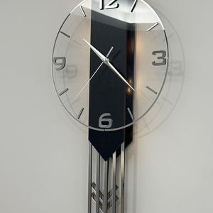 May include: Modern wall clock with a clear glass face and silver hands. The clock has a black rectangular backing and a decorative silver pendulum. The numbers are silver and easy to read. The clock is mounted on a light gray wall.