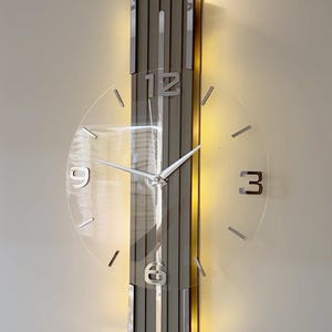 May include: A vertical wall clock featuring a central grey panel with chrome trim and integrated backlighting. A circular transparent face overlays the panel, displaying silver numerals at 12, 3, 6, and 9, along with silver hour markers and hands.