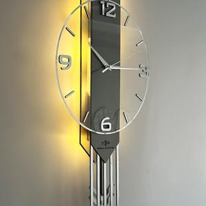 Pode incluir: Relógio de parede moderno com mostrador transparente e ponteiros prateados. O relógio tem um fundo cinza com um design vertical e uma luz atrás. Os números 12, 3, 6 e 9 são visíveis. O relógio está rotulado como "WALL CLOCK".