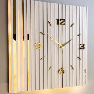 May include: A rectangular wall clock featuring a white slatted surface with vertical gold accents. The clock face displays gold-toned numerals at the 12, 3, 6, and 9 positions, with thin gold hands and simple gold markers for the remaining hours.