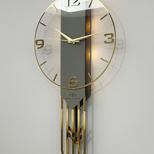 May include: A modern wall clock with a clear glass face and gold-colored hands and numerals. The clock has a gray backing and a decorative gold pendulum. The text "WALL CLOCK" is visible on the clock face.