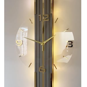 May include: A wall clock featuring a vertical, grey, panelled rectangular centre with gold trim. A transparent circular face overlays the centre, displaying gold-coloured numbers 3, 6, 9, and 12, along with thin gold hour markers and gold clock hands.