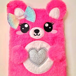 May include: A bright pink plush notebook cover shaped like a bear with fuzzy fur. It features a large iridescent glitter bow on one ear, embroidered black eyes with long lashes, a white muzzle, and a central white patch with a silver glitter heart.