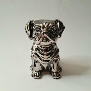 May include: A reflective, metallic silver ceramic figurine of a seated pug dog. The sculpture features a smooth, high-gloss finish that captures light, highlighting the detailed facial wrinkles, floppy ears, and the distinct, sturdy posture of the dog.
