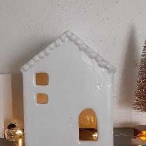 May include: A white ceramic house-shaped luminary featuring a gabled roof with a beaded edge, two small square windows on the left side, and a large arched doorway on the right. The interior is illuminated by warm light from a string of fairy lights.