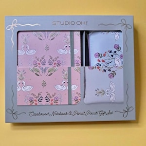 Puede incluir: Un juego de regalo en una caja gris que presenta un cuaderno encuadernado, un estuche para lápices y un cuaderno pequeño. El cuaderno y la bolsa tienen un fondo rosa con cisnes blancos y diseños florales. La caja dice "STUDIO OH!" y "Casebound Notebook & Pencil Pouch Gift Set."