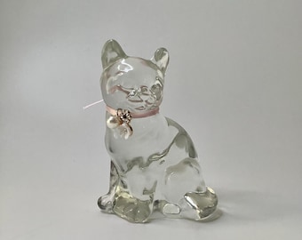 May include: A clear glass cat figurine in a seated position. The cat has a pink ribbon collar with a decorative charm and a pearl. The figurine is transparent and sits on a white surface.