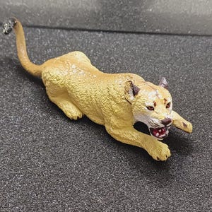 May include: A tan cougar figurine in a stalking pose, with its mouth open, revealing teeth. The figurine has a textured surface and is set against a dark background. The tail is curved upwards.