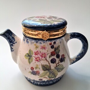 May include: A ceramic teapot-shaped trinket box featuring a hand-painted blackberry and pink flower motif. The piece has a dark blue sponged rim, a gold-toned metal hinged closure with a floral clasp, and a matching lid with a blackberry design.