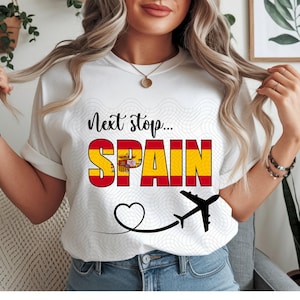 May include: White t-shirt with the text "Next stop... SPAIN" in black and red, with the Spanish flag incorporated into the word "SPAIN". A black airplane graphic and a heart-shaped trail complete the design. The shirt is a casual travel-themed item.