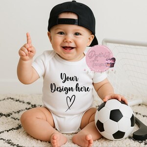 May include: A baby wearing a white onesie and a black baseball cap, smiling and pointing. The onesie has the text "Your Design here" and a heart. A black and white soccer ball is held in the baby's hand.