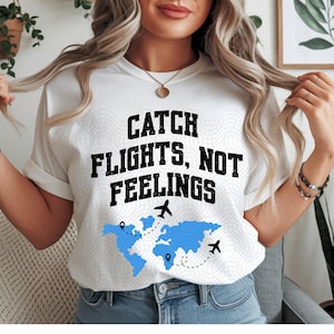 Op de afbeelding: Wit T-shirt met de tekst "CATCH FLIGHTS, NOT FEELINGS" in zwarte letters. Onder de tekst staat een blauwe wereldkaart met vliegtuigafbeeldingen en stippellijnen. Het shirt heeft een casual stijl.