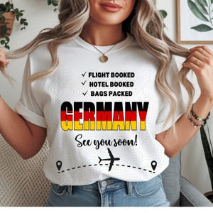 Może przedstawiać: Biała koszulka z napisem "GERMANY" w kolorach flagi Niemiec. Koszulka zawiera również napisy "FLIGHT BOOKED", "HOTEL BOOKED" i "BAGS PACKED". Poniżej napis "See you soon!" z grafiką samolotu.