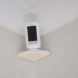 May include: White Ring security camera mounted in a corner. The camera has a black lens and a speaker grill. The camera is mounted on a white triangular base.