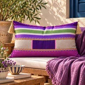 Puede incluir: Un cojín rectangular con rayas horizontales en morado, verde y blanco, un panel central de bloque morado, secciones laterales beige, ribetes decorativos amarillos y borlas de color canela claro en las esquinas superiores del cojín.