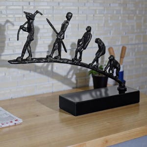 May include: A black metal sculpture depicting the evolution of man, from ape to human, mounted on a black rectangular base. The sculpture is placed on a wooden table with a brick wall background.