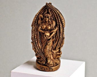 May include: A bronze-colored statue of Lilith, a female figure with horns, wings, and snakes. She holds a skull and wears a crown. The statue is on a white surface and has the word "LILITH" at the base.