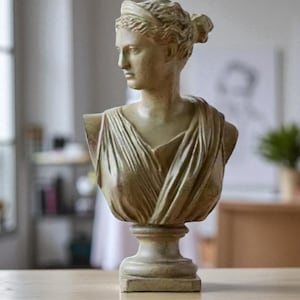 May include: A classical-style bust sculpture in a muted beige tone. The sculpture depicts a woman with her hair pulled back, wearing draped garments. The bust is mounted on a pedestal base, and is set on a light-colored wooden surface.