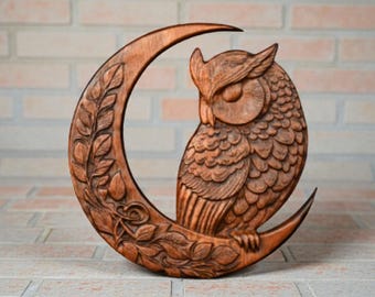 May include: A wooden sculpture of an owl perched on a crescent moon. The owl is intricately carved with detailed feathers and facial features. The moon is decorated with leaf and floral patterns. The sculpture is a warm brown color.
