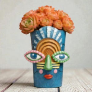 Pode incluir: Um vaso azul pintado à mão com um design de rosto, segurando um cacho de flores laranja. O vaso apresenta olhos decorativos, um nariz e uma boca. O design é uma reminiscência da arte tribal.
