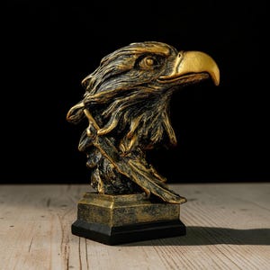 Puede incluir: Un busto esculpido de una cabeza de águila con un acabado metálico en oro y negro. El águila se muestra de perfil, mirando hacia la derecha, con texturas de plumas detalladas y un pico curvo prominente sobre una base cuadrada negra.